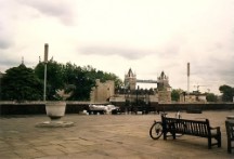 A bum wrapped in plastic sleeping on a bench with the London Tower Bridge, England in the background. Ted's bike is resting against the bench.